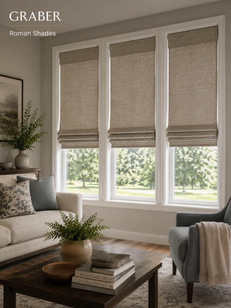 A bright living room features three large windows with beige Graber Roman shades, a gray armchair, a light sofa, a wooden coffee table with a potted plant, and greenery outside. Graber Roman Shades logo is in the top left.