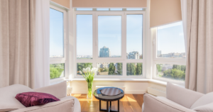 Bright living room with large corner windows fitted with sleek blinds, two white armchairs, a small round table, a vase of white tulips, and a cityscape view with modern buildings and greenery outside.